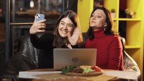 Two beautiful young women in a bar are chatting via video call on a smartphone, smiling happily, and waving