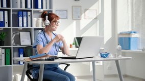 young female doctor in glasses and a headset works at a laptop and communicates with a patient via video call while sitting in a medical office