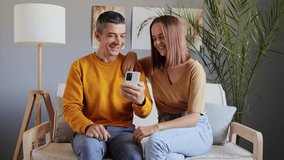 Home phone. Wireless connection. Smartphone happiness. Love and technology. Cheerful adult couple using phone at home while sitting on sofa watching funny video laughing happily.