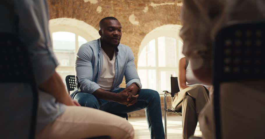 A Black man in a blue shirt expresses his thoughts in group therapy while other participants applaud him at a group therapy meeting in a yellow brick hall Royalty-Free Stock Footage #1109670709