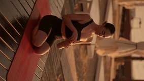 Vertical video: A brunette girl in a black sports summer uniform sits on a red mat and drinks water from a sports bottle after doing yoga on a sunny beach