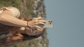 Vertical video. At sunrise, a young woman captures the mountain views on her phone while squinting from the bright sun.