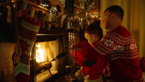 Dad and son together by cozy fireplace with hanging stockings and Christmas garland lights. Family at Winter and Christmas holidays. Young father with cute boy using bellows to blow air into fireplace Arkistovideo