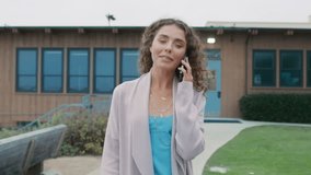 Laughing curly woman wearing trendy boho style cardigan and blue silk dress walks by green park and uses smartphone. Pretty happy woman walks at ocean talking by mobile phone on cloudy summer day 4K