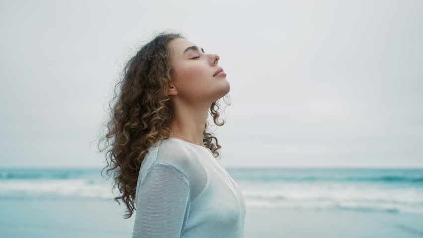 Calm young woman taking deep breath of fresh air meditating with eyes closed standing outdoors at ocean waves on background. Side profile view girl enjoy moment of peace, mental relaxation, no stress Royalty-Free Stock Footage #1109672975