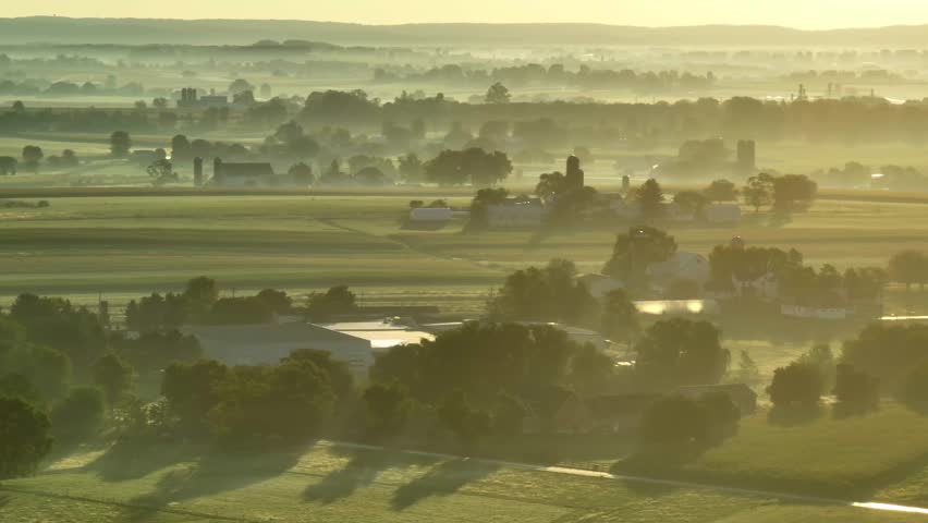 Fog, mist, and dew over rural farmland in USA during morning sunrise. Aerial establishing shot of corn fields, farms, barns, silos, and trees.