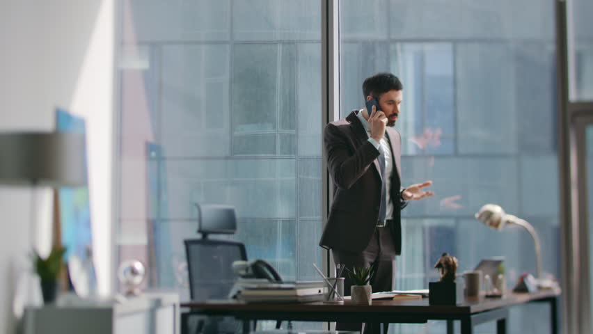 Financial manager speaking mobile phone standing modern workplace looking notes on work desk. Busy bearded entrepreneur calling subordinates discussing issues. Serious boss communicating by smartphone
