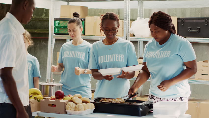 Multiracial volunteer ladies serve meals and non-perishable products to hungry and homeless people. Friendly charity workers handing out free food to the poor and needy. Tripod shot. Royalty-Free Stock Footage #1109680795