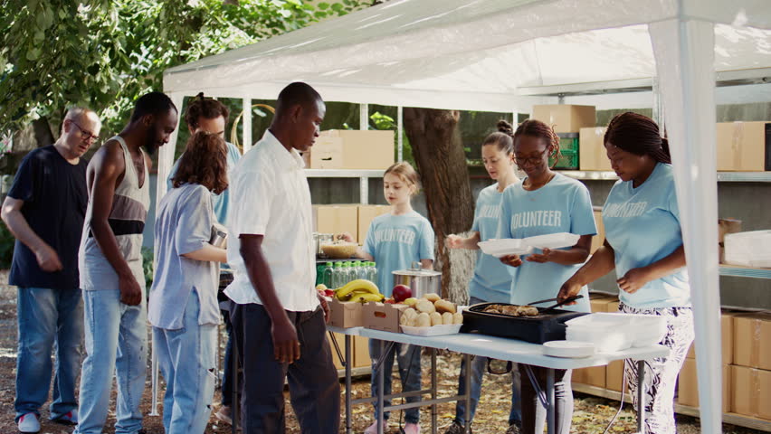 People from many backgrounds assemble outside to distribute non-perishable donations and help the poor and homeless. Multiracial volunteers provide needy individuals with free, freshly prepared meals. Royalty-Free Stock Footage #1109680809