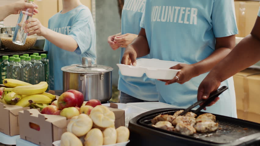 Multiethnic people in blue t-shirts, assembled outside to hand out food donations and help the homeless and hungry. Focus on volunteers serving free, freshly prepared meals to needy individuals. Royalty-Free Stock Footage #1109680853