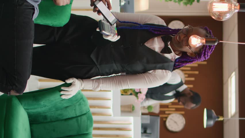 Vertical video Traveller paying for coffee cup to hotel staff, using nfc mobile payment at pos while he waits for room registration. African american client buying drink, discussing with receptionist.