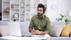 Confident coach in headset and glasses talking on video call using laptop sitting on sofa in living room at home office. Young tutor conducts an online training, seminar, course or webinar remotely