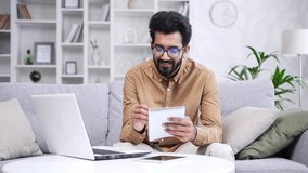 Young handsome bearded male student listens to online e-learning using laptop while sitting in home office. Employee on remote advanced training courses writes in a notebook communicates with a tutor