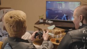Back view of boy playing FPS video game with controller while his girlfriend feeding him with pizza, sitting on couch together with friends on weekend night