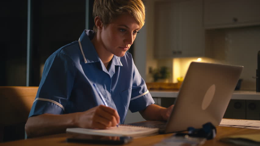 Tired woman wearing nurse's uniform working or studying on laptop at home at night drinking hot drink - shot in slow motion Royalty-Free Stock Footage #1109690911
