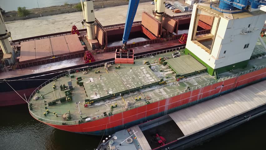 Aerial drone footage of a bulk carrier cargo ship being unloaded by a crane at a port. Bulk cargo tanker loading at a port seen from a bird's eye view. Royalty-Free Stock Footage #1109690995