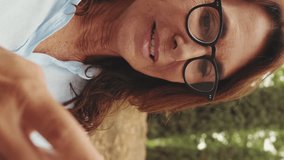 Vertical video, Close-up of beautiful retired woman in glasses using phone while sitting in park
