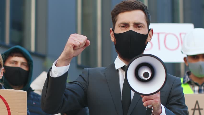 Portrait of male activist in suit and tie speaking, screaming in megaphone speaker. Man participate in anti quarantine protest. Multiethnic group of young people demonstrating against lockdown. Royalty-Free Stock Footage #1109694493