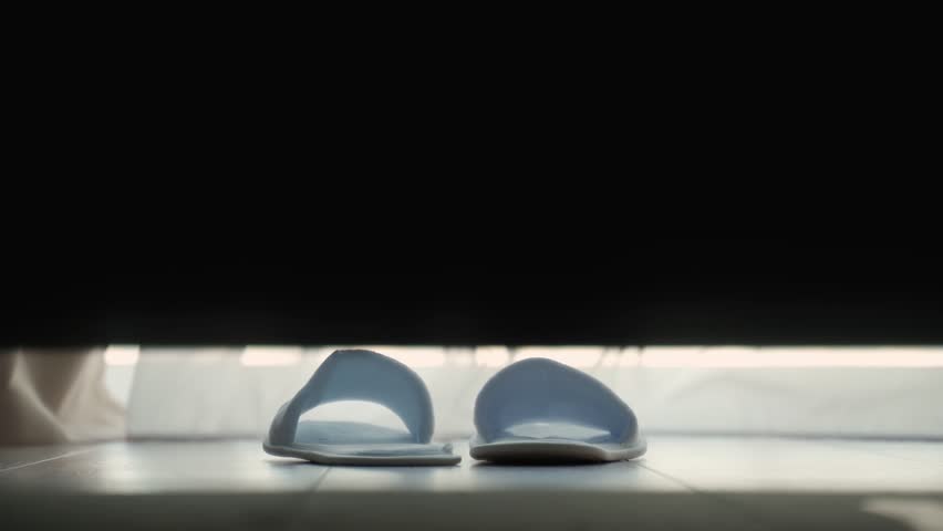Woman puts on white slippers, getting out of bed on the floor in a hotel room. The guest puts her feet in slippers gets out of bed and leaves close-up. 
