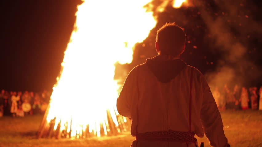 Person in Slavic clothes worshipping big fire at night. Ethnic holiday Ivan Kupala in village.