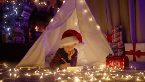 Funny kid in large red Santa hat comfortably lying in tent with magic light garland. Cute little boy writing wishes in letter to Santa Claus in fairy tale atmosphere in living room with fireplace 4K - Powered by Shutterstock - Get 15% off with code: PIKWIZARD15