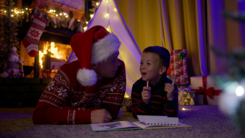 Caring father in red Santa Claus hat having fun time together with cute little son before going to bed, lying on floor under teepee tent decorated with lights in decorated room with burning fireplace Royalty-Free Stock Footage #1109726181