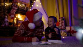 Caring father in red Santa Claus hat having fun time together with cute little son before going to bed, lying on floor under teepee tent decorated with lights in decorated room with burning fireplace - Powered by Shutterstock - Get 15% off with code: PIKWIZARD15