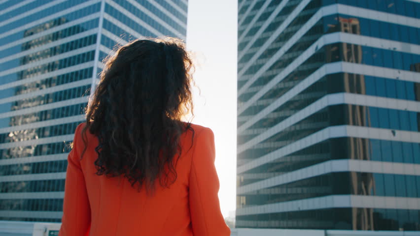 Low angle back view of beautiful brunette sexy woman with gorgeous curly long hair flying on wind in cinematic golden sunshine. Smiling woman portrait turning and looking to camera over shoulder 4K