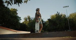 Bottom-up shot of a blonde girl in a plaid shirt and shorts standing majestically with a skateboard in a skate park in summer