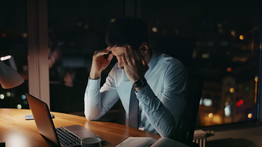 Frustrated professional employee working late evening in dark office. Overloaded businessman looking laptop monitor feeling headache from overwork. Troubled manager tired of deadline at night close up