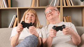 Happy elderly couple sitting on the sofa at home, they are playing games holding joysticks in their hands