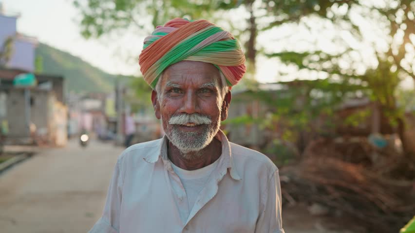 An Indian ethnic happy man or male is wearing a traditional turban or headwear facing or looking at the camera and smiling while standing outdoors against the rising sun in a village Royalty-Free Stock Footage #1109739993