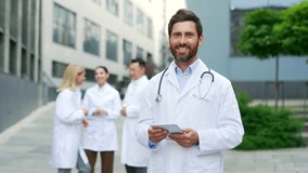 Portrait of a confident happy male doctor looks at camera. Sіmiling positive mature bearded male medical physician, medic stands outside a hospital or clinic building holding a tablet outdoors  - Powered by Shutterstock - Get 15% off with code: PIKWIZARD15