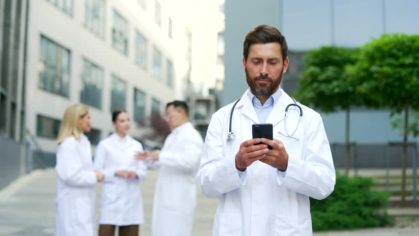 Mature adult male doctor using mobile phone while walking near clinic or hospital office. Male physician going browsing chatting typing text messages reading smartphone. Outdoors outside Royalty-Free Stock Footage #1109740643