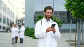 Mature adult male doctor using mobile phone while walking near clinic or hospital office. Male physician going browsing chatting typing text messages reading smartphone. Outdoors outside - Powered by Shutterstock - Get 15% off with code: PIKWIZARD15