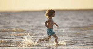 Happy smiling kid 5-7 years old running along beach in water slow motion. Boy with long hair in blue swimming trunks runs cheerfully along beach in setting sun. Concept beach holiday for children