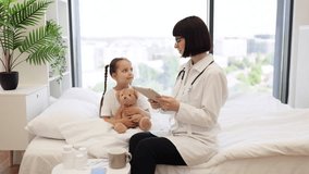 Experienced female pediatrician typing on digital tablet detailed info about little sick patient during home visit. Tired caucasian girl staying in bed in embrace with her plush teddy bear. - Powered by Shutterstock - Get 15% off with code: PIKWIZARD15