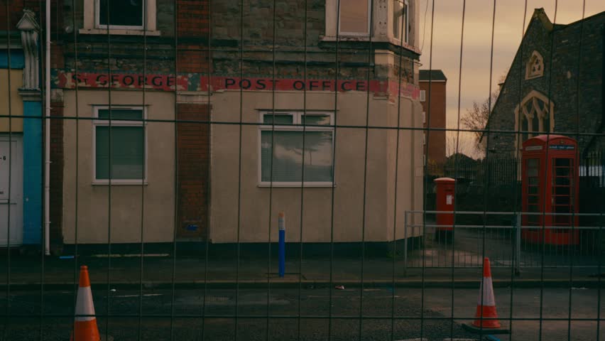 Disused, derelict post office behind wire fence at sunset. Church in the background.