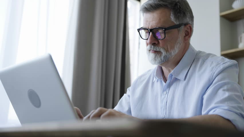 Older man working online with laptop computer at home sitting at desk. Home office, surfing internet | Shutterstock HD Video #1109744633