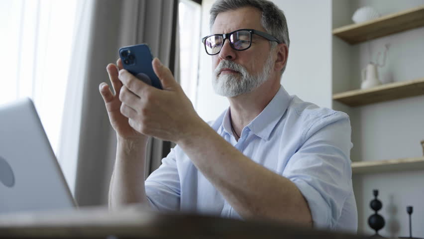 Professional Mature Man Sitting at His Desk in Home Office Working on a Laptop. Man Using Smartphone to Check Social Media, Search Internet | Shutterstock HD Video #1109744677