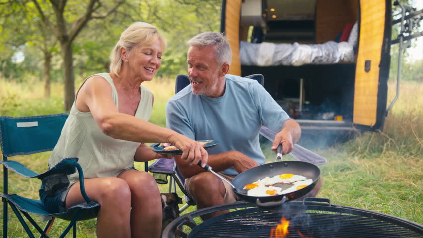 Senior retired couple camping in countryside in RV cooking eggs for breakfast in frying pan outdoors on fire - shot in slow motion