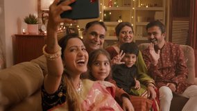 Happy smiling Hindu ethnic Indian family members in traditional attire posing to take or click a selfie picture or photo together using a mobile phone or smartphone during Diwali festival season.