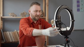 bearded Caucasian male archaeologist blogger is recording the broadcast in his studio using a smartphone and a ring lamp. shows an old vase, takes it apart and wipes the dust off it