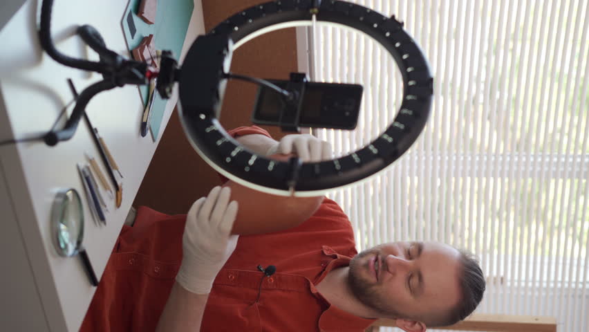 a bearded Caucasian male archaeologist blogger is recording the broadcast in his studio using a smartphone and a ring lamp. shows an old vase being dismantled.