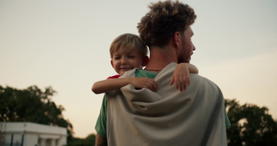 A little blond boy lovingly hugs the neck of his dad with curly hair in a green T-shirt. Little boy loves his dad and hugs him Royalty-Free Stock Footage #1109759089