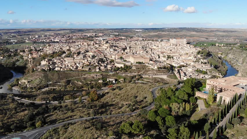 Aerial crane shot of Toledo, Spain. Partially cloudy. Historical and medieval old town with cathedral, towers, and Alcazar. River flowing around city.