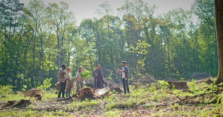 Multiage and multicultural group of enthusiastic attractive people doing forestation work in woods helping nature. Adults and children volunteering plating trees listening to instructions.