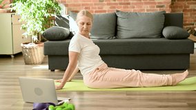 Senior woman on yoga mat doing yoga pose and watching online classes on laptop, workout in living room. Active lifestyle concept