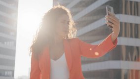 Happy bi racial diverse woman taking selfie, taking selfie and blowing kiss, posting on social media at sunset. Fashion lifestyle influencer blogger using mobile smartphone in cinematic sun lens flare