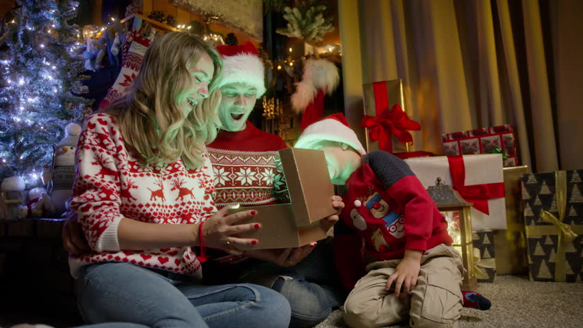 Happy young parents receiving golden gift box from little loving son in red Santa hat, expressing gratitude for pleasant surprise and attention at Christmas celebration. Winter holidays congratulation